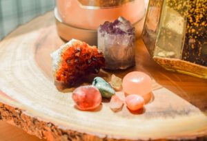 crystals sitting on a natural wood table. crystals range in color from pink to purple and orange. they are various sizes from the size of small 1 or 2 inch rock to the size of a small pillar candle.